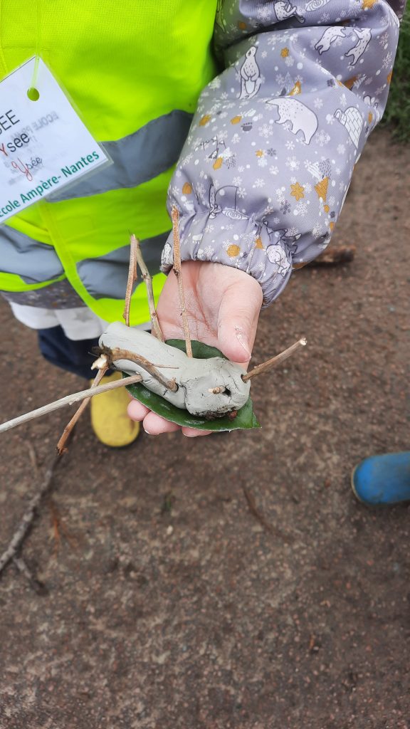 un enfant tient dans sa main une petite bête en argile crée avec des brindilles pour les pattes et les antennes sur une feuille d'arbre