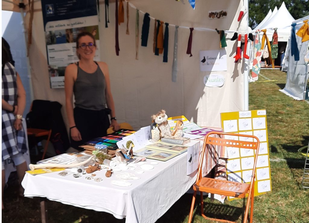 un stand avec une table et l'animatrice. Sur la table, on voit des indices de présence d'animaux comme des pommes de pin rongées, un bois de cerfs, et des photos d'oiseaux